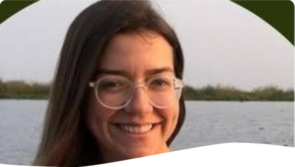 virginia is a white woman, with shoulder-length brown hair, and wears prescription glasses. It's smiling and we can see a river in the background.
